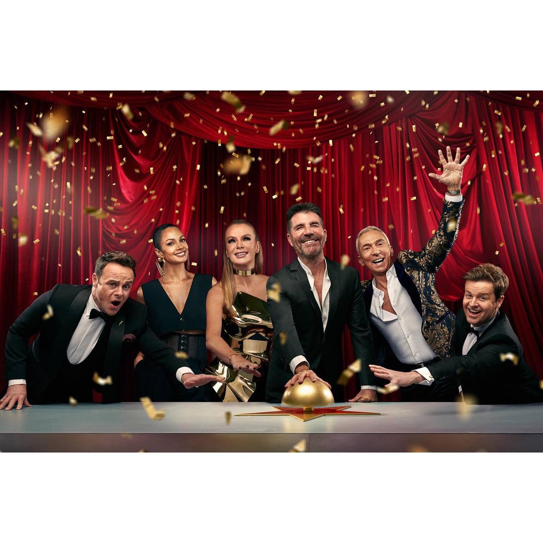 Group shot of six judges or hosts in formal attire with confetti falling in front of a red curtain, posing behind a table with a gold button.