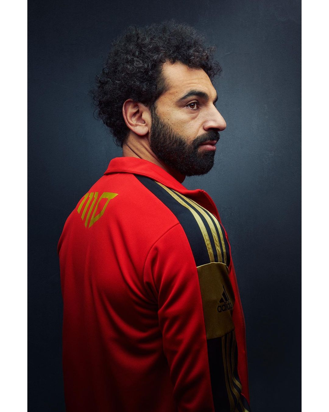 Portrait of adult male with a beard and curly hair in red Adidas tracksuit, looking to the side against a dark backdrop in a close-up shot.