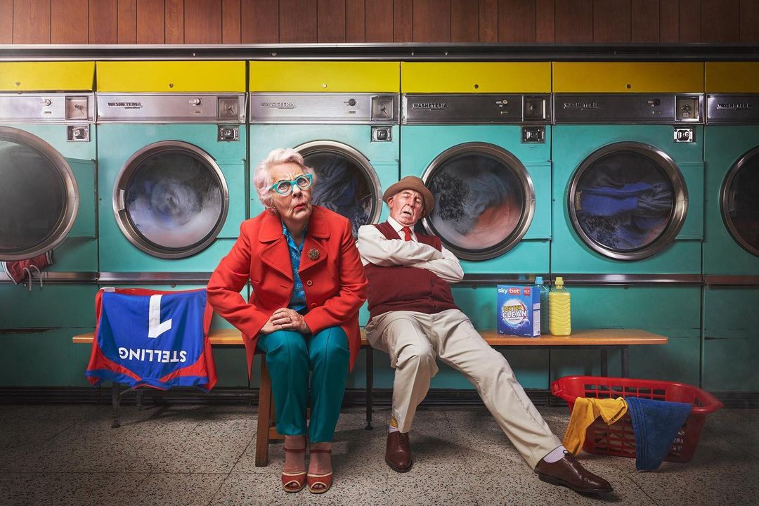 Two elderly people sitting in a laundromat with multiple washing machines in the background, shirts, detergent, and laundry basket.