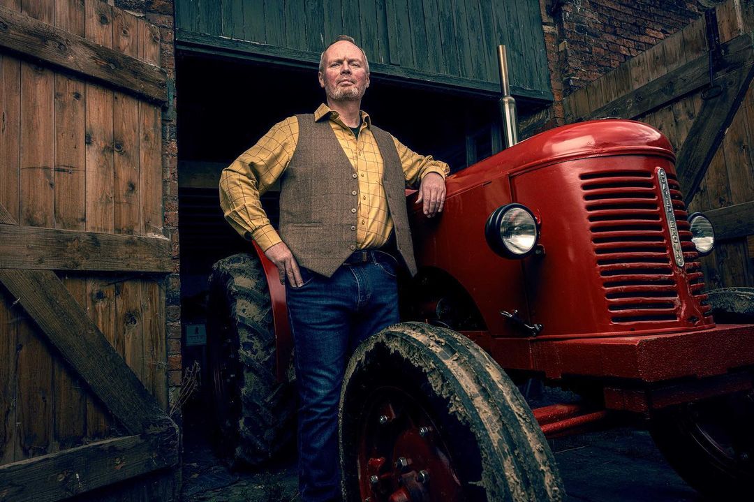 Adult male poses next to a red tractor with open barn doors and brick walls visible in the background. He wears jeans and a vest.