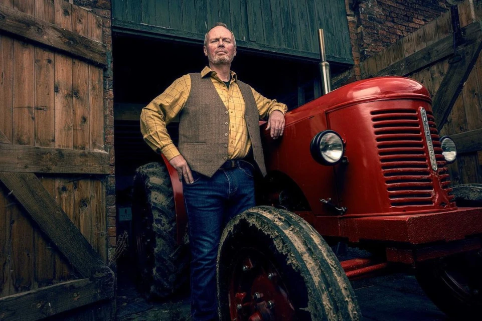 Adult male poses next to a red tractor with open barn doors and brick walls visible in the background. He wears jeans and a vest.