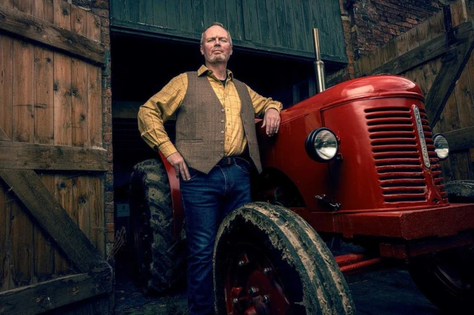 Adult male poses next to a red tractor with open barn doors and brick walls visible in the background. He wears jeans and a vest.