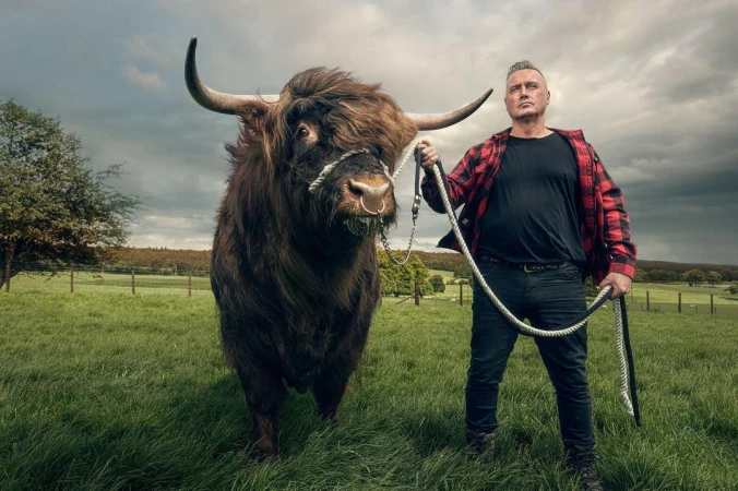 A man in a flannel shirt stands in a green field holding a Highland bull with large horns and long shaggy hair, under a cloudy sky.