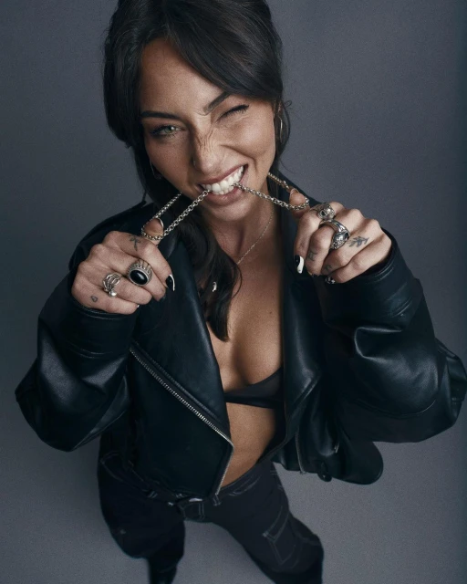 Eye-level shot of a stylish woman in a black leather jacket biting a silver chain necklace, rings, and long black nails against grey background.