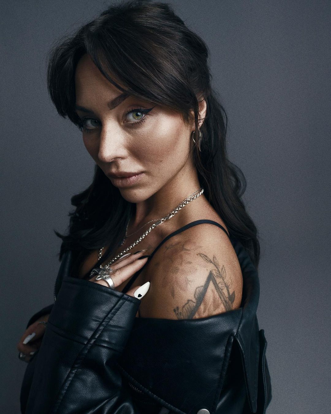 Portrait of a woman wearing a black leather jacket with visible tattoos and silver jewelry against a grey backdrop.