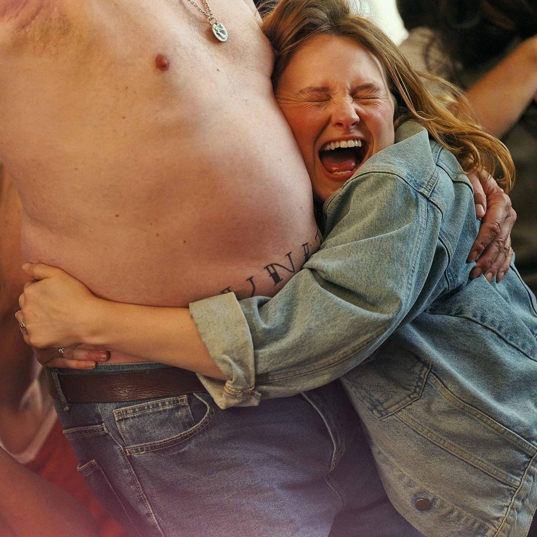 A joyful young woman hugs a shirtless man, her face expressing elation, set against a soft-focus background.