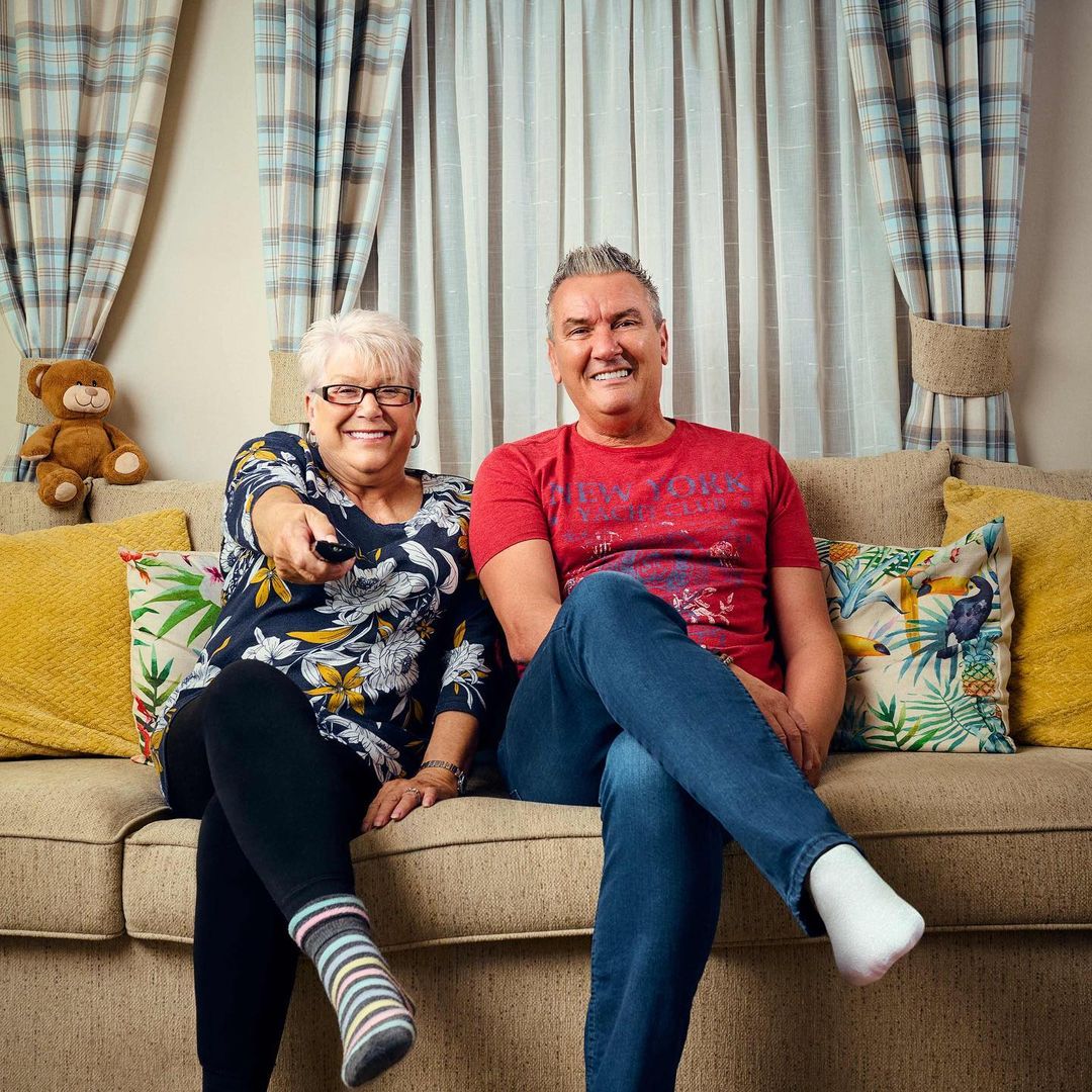 An elderly couple relaxes on a beige sofa, watching TV with a remote in hand, set against patterned curtains and decorative pillows.