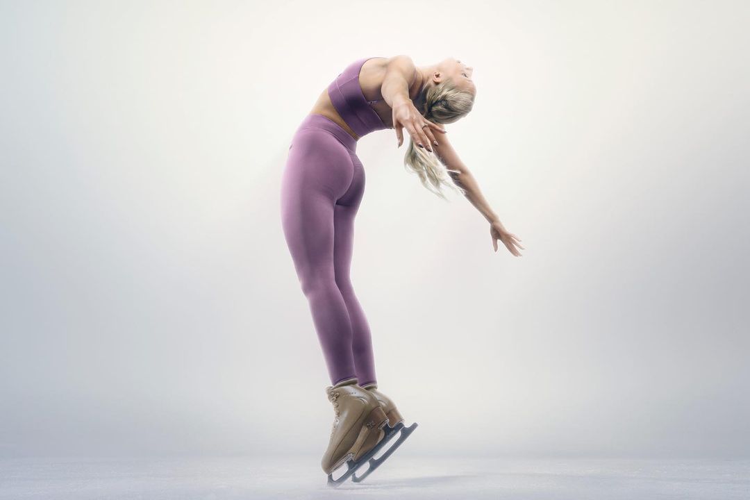 Female figure skater gracefully arches backward on ice, showcasing athletic skill in sportswear against a soft, diffused background.