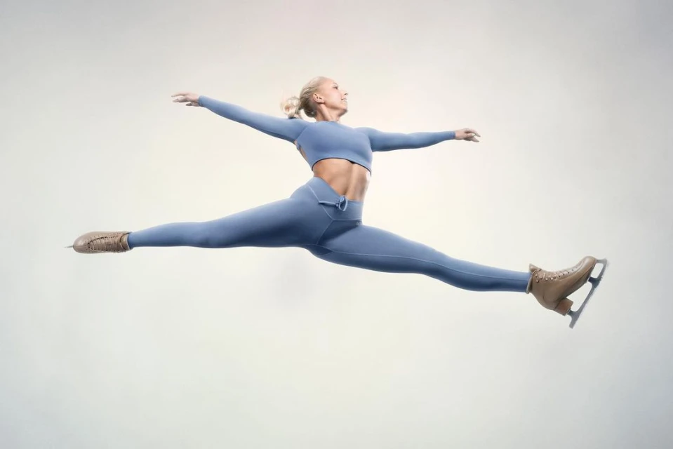 Female figure skater leaps mid-air, performing a split with arms extended, wearing a blue athletic outfit and brown ice skates, on white backdrop.