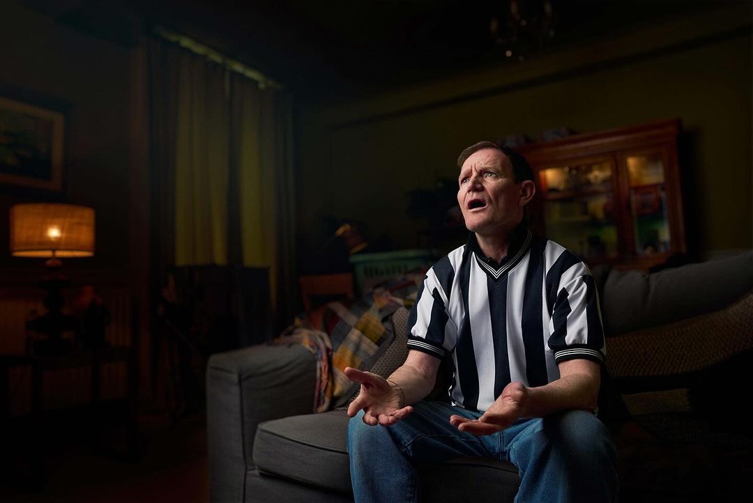 Adult man wearing a black and white striped v-neck shirt sits on a grey couch indoors with an emotional expression, looking upward.