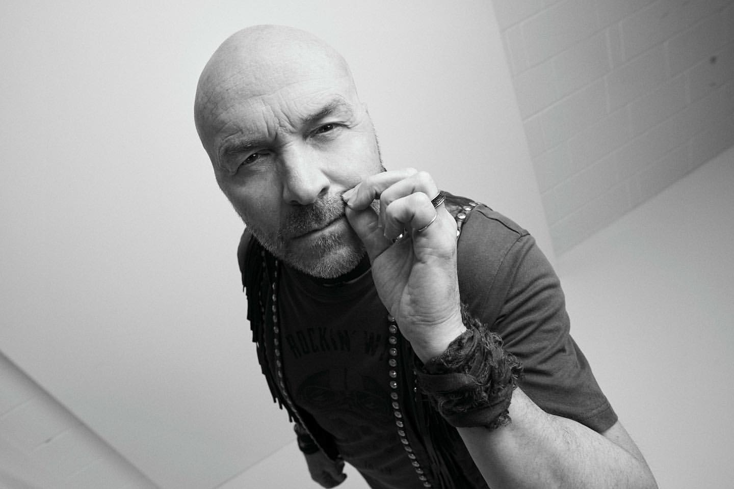 Monochrome portrait of a bald adult male, viewed from a low angle, wearing a studded vest and t-shirt, with hand near his mouth.