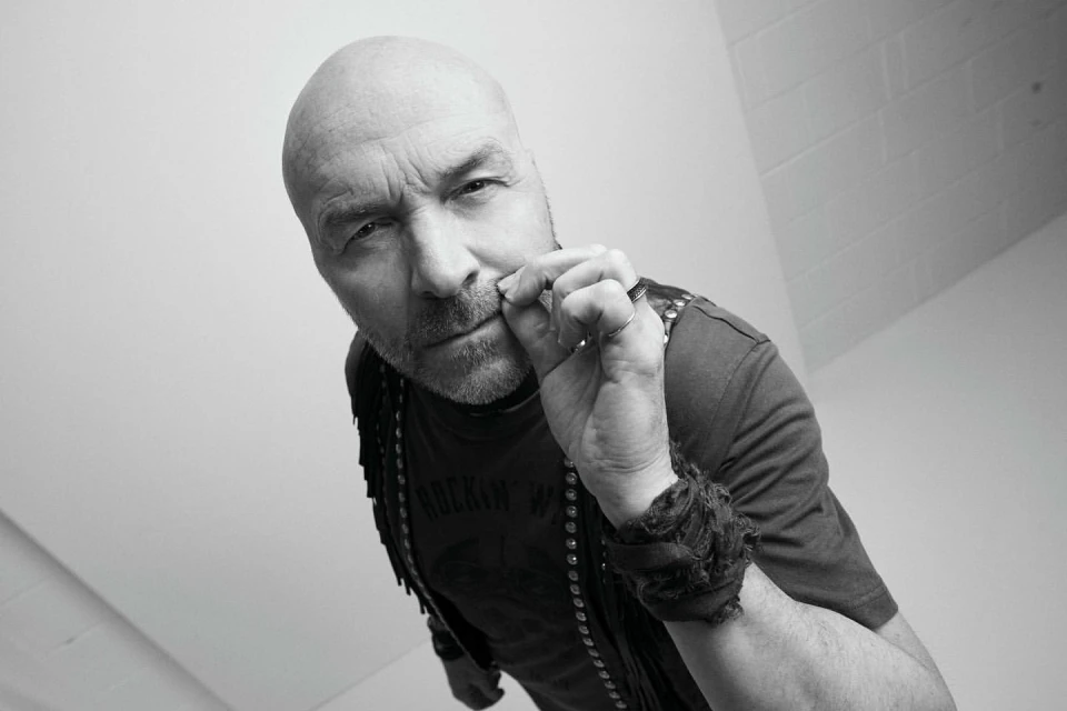 Monochrome portrait of a bald adult male, viewed from a low angle, wearing a studded vest and t-shirt, with hand near his mouth.