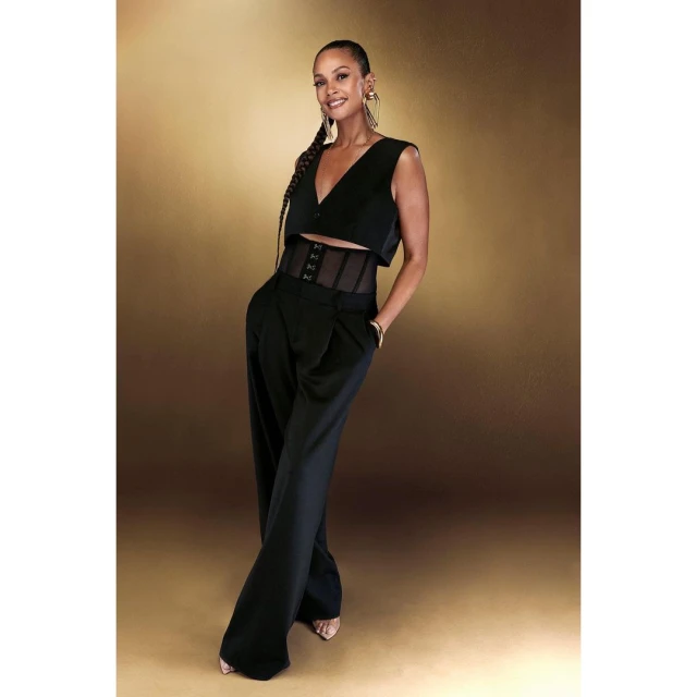 Full-length studio shot of an adult woman with a long braid, wearing a black two-piece outfit, posing on a bronze background.