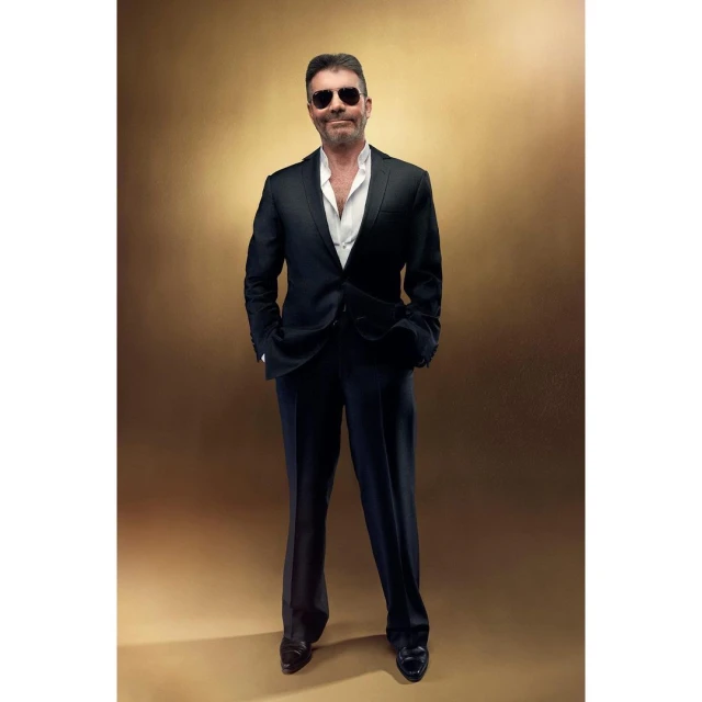 Full-length portrait of an adult male with sunglasses wearing a black suit with a white shirt on a bronze background indoors.