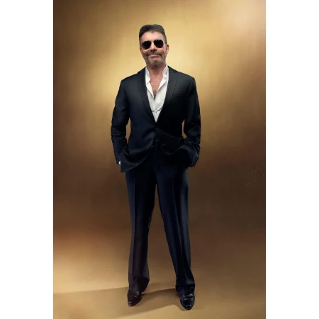 Full-length portrait of an adult male with sunglasses wearing a black suit with a white shirt on a bronze background indoors.