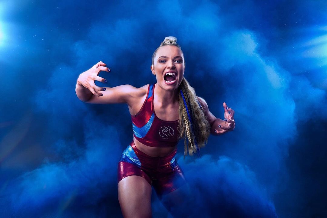 Action shot of a blonde woman in a red and blue sports outfit, roaring with hands raised against a blue smoky background.
