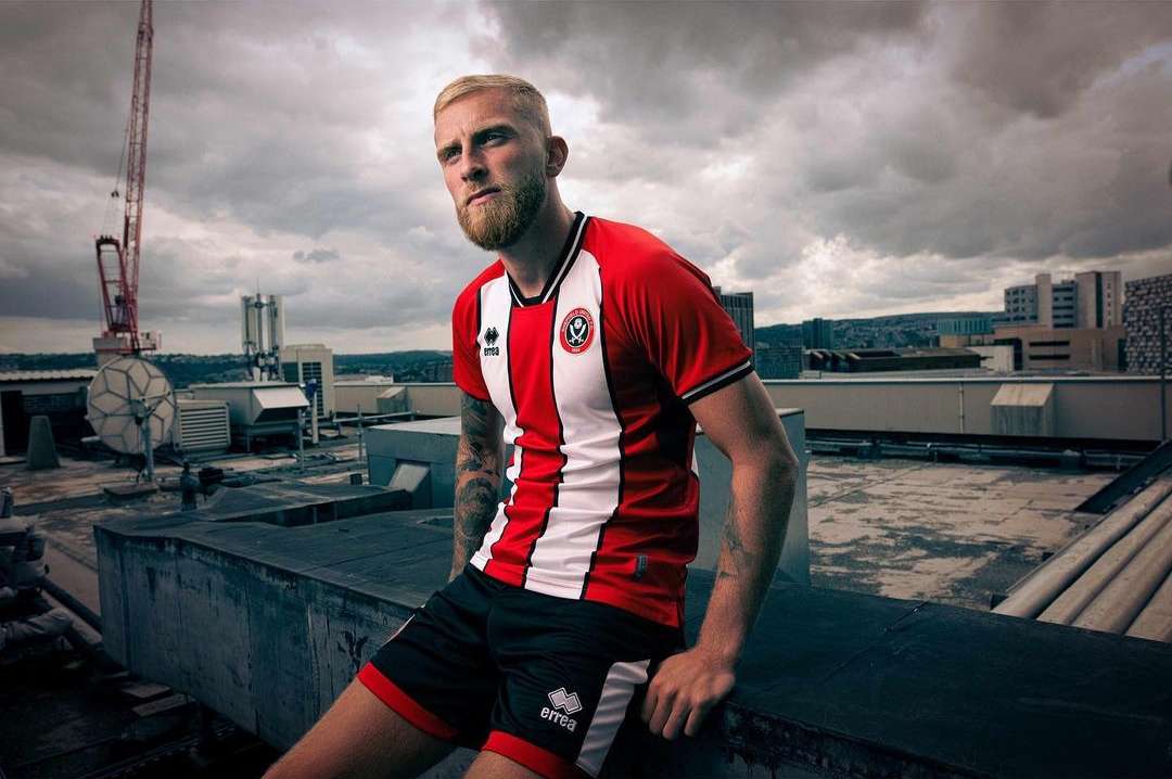 Athletic male model in Sheffield United soccer uniform posing on rooftop with city view and cloudy sky background, urban setting, eye-level.