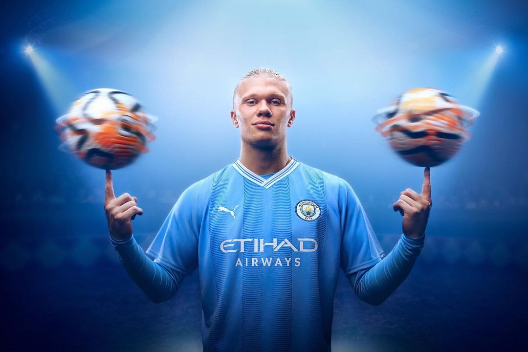 Soccer player in a light blue jersey balancing two soccer balls on his fingers in a promotional studio shot with spot lights.
