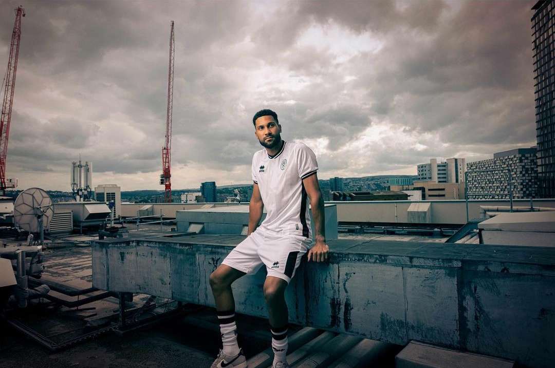 A man in a sports uniform sits on a concrete rooftop against a cloudy skyline with construction cranes and city buildings in the distance.