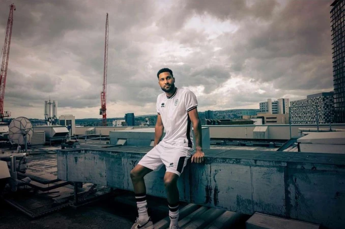 A man in a sports uniform sits on a concrete rooftop against a cloudy skyline with construction cranes and city buildings in the distance.