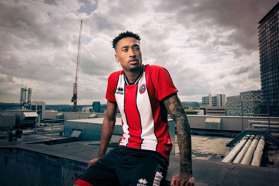 Adult male athlete in a red and white sports jersey and dark shorts sits on a rooftop overlooking a city skyline with cloudy skies.