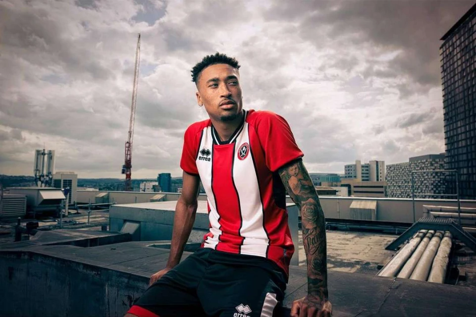 Adult male athlete in a red and white sports jersey and dark shorts sits on a rooftop overlooking a city skyline with cloudy skies.