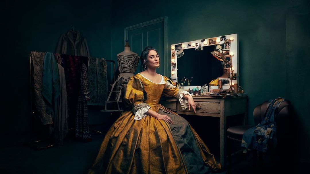 A woman in a golden gown sits in front of a vanity table in a dimly lit room with a mannequin and clothing rack.
