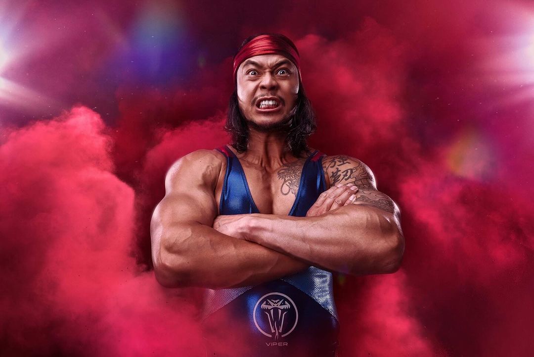 Muscular man with determined expression and arm tattoos wearing red headscarf and blue wrestling leotard in smoky atmosphere.