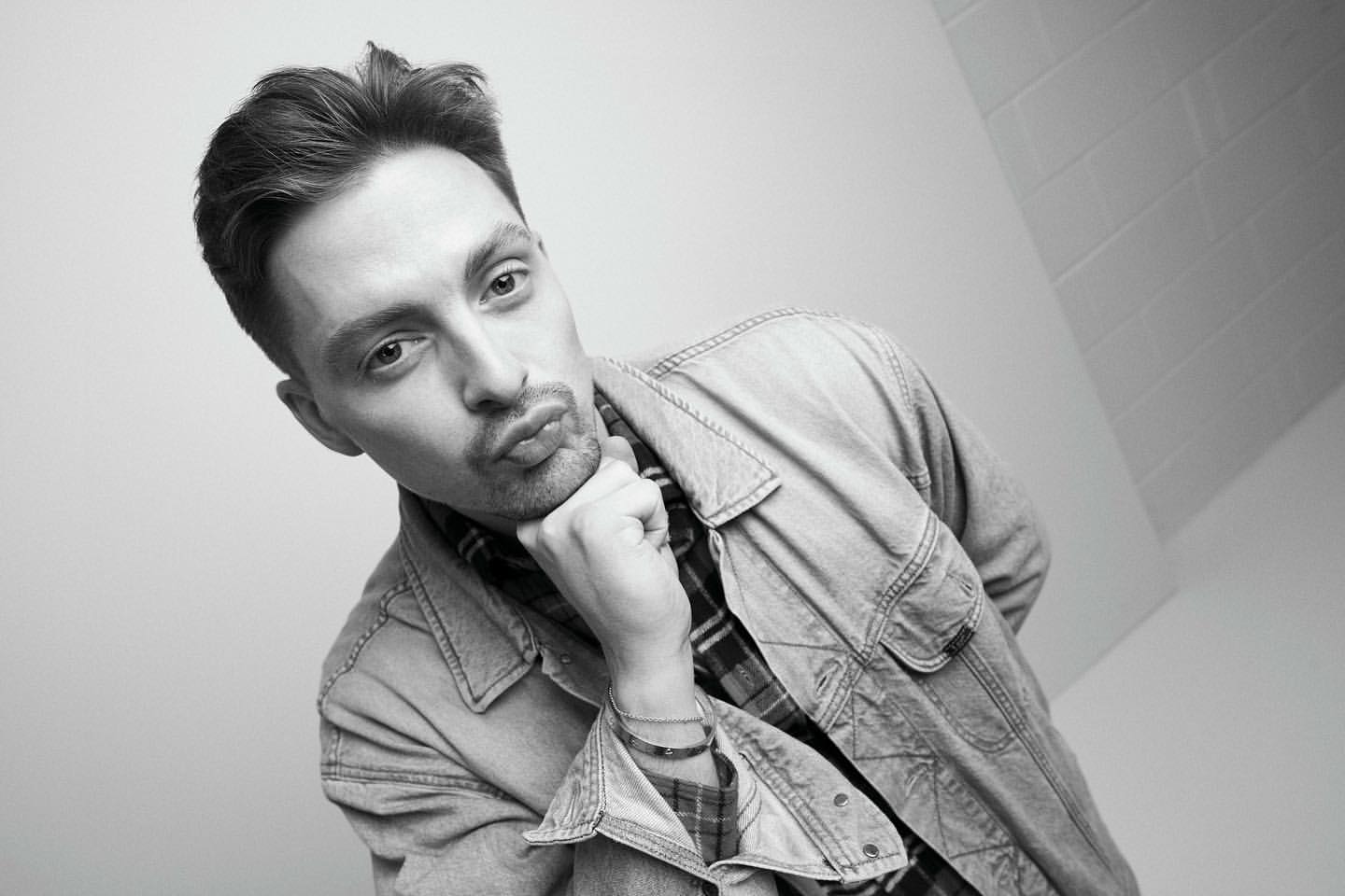 Black and white portrait of a young adult male with styled hair, wearing a denim jacket and making a kissing face in a studio setting.