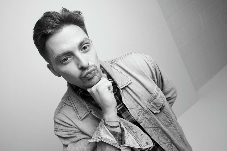 Black and white portrait of a young adult male with styled hair, wearing a denim jacket and making a kissing face in a studio setting.