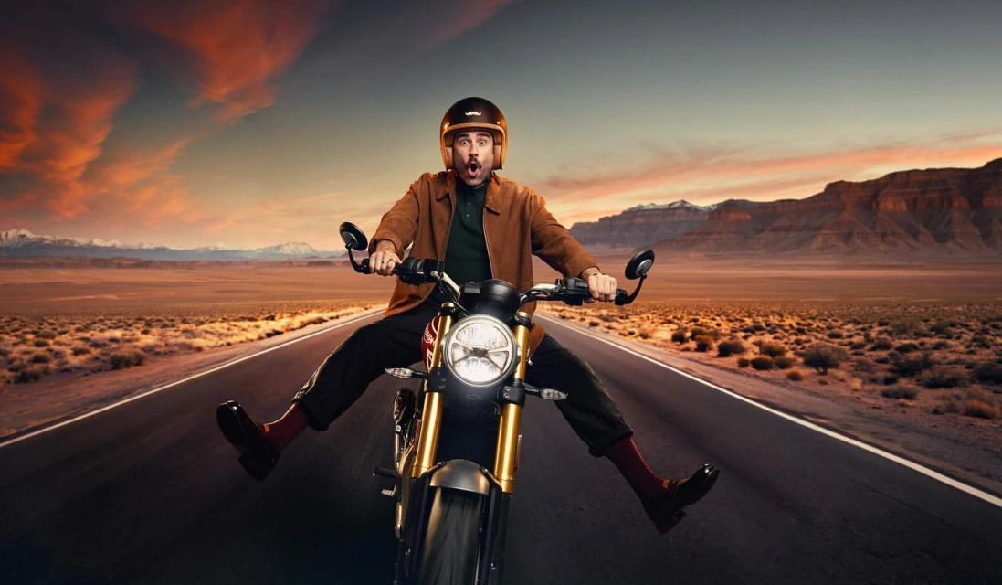 Adult male with a surprised expression is riding a yellow motorcycle with no hands along a desert road at sunset, with mountains.