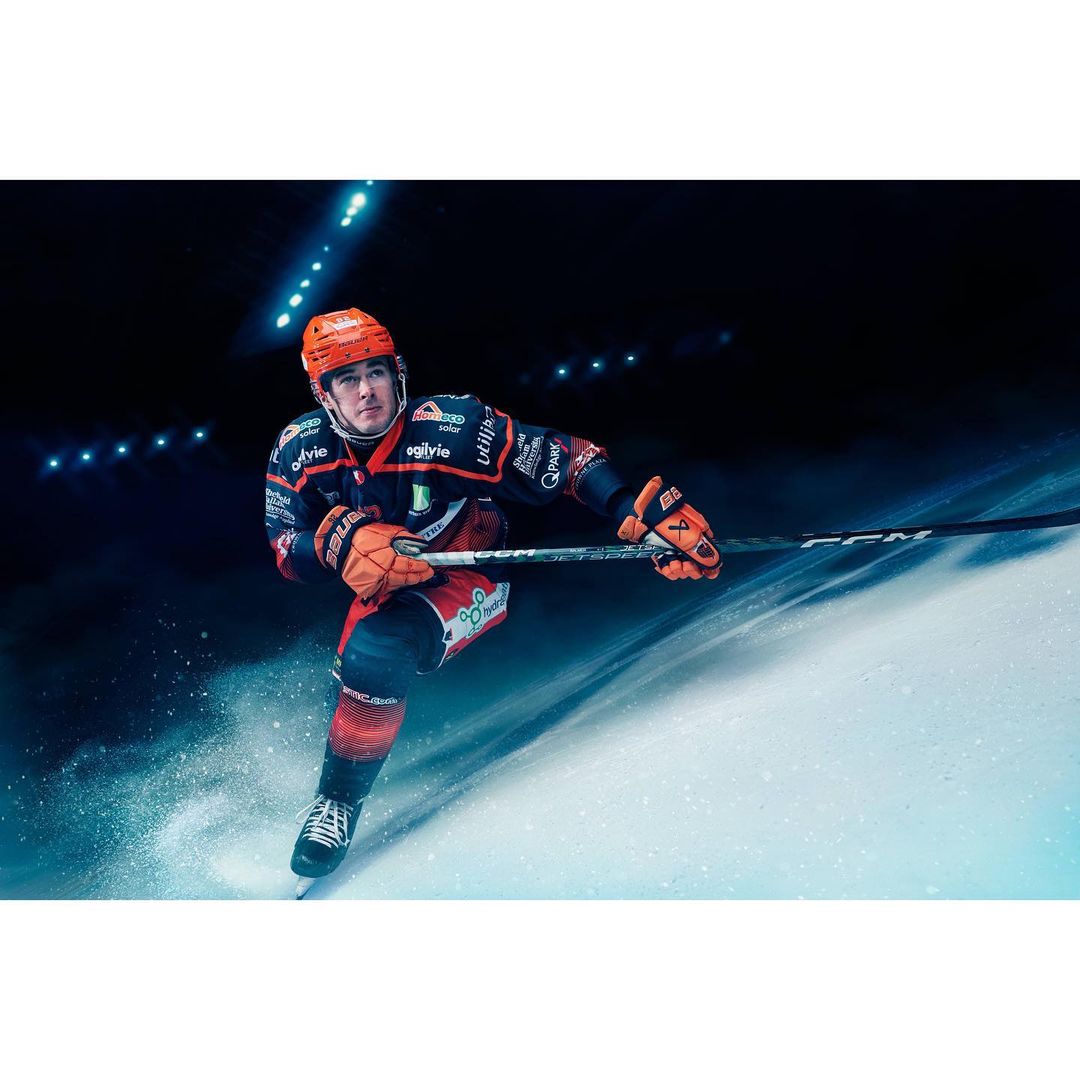 Hockey player in full uniform skates on ice, wearing a branded jersey, helmet and gloves, with a hockey stick in hand in an arena.