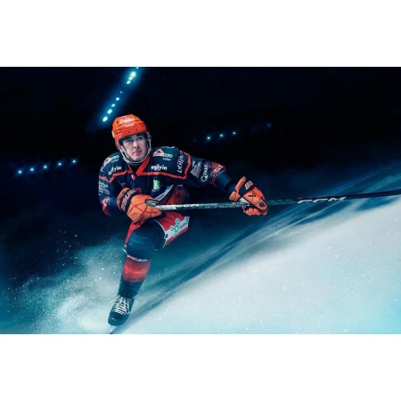 Hockey player in full uniform skates on ice, wearing a branded jersey, helmet and gloves, with a hockey stick in hand in an arena.