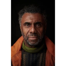 Portrait of an older Black man with a graying beard wearing a patterned sweater, olive scarf, and burnt orange jacket against a dark background.
