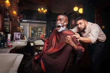 Black man with shaving foam on his face reacting with wide-eyed surprise as a barber holds a straight razor in a traditional barbershop