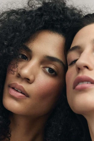 Close-up portrait of two women with dark curly hair posing closely together with natural makeup.