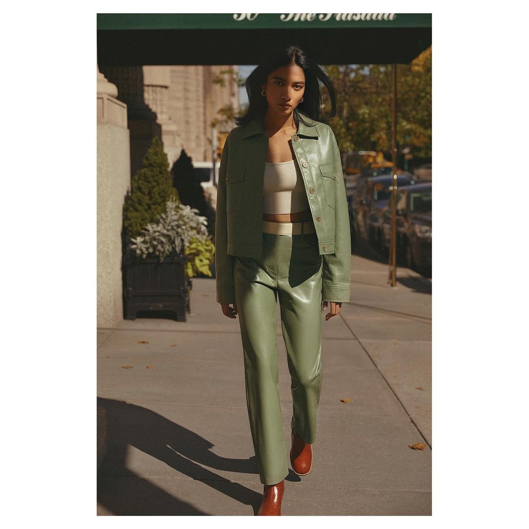 Woman in light green leather jacket and pants poses on urban sidewalk near building, showcasing modern fashion with a neutral top and tan boots.