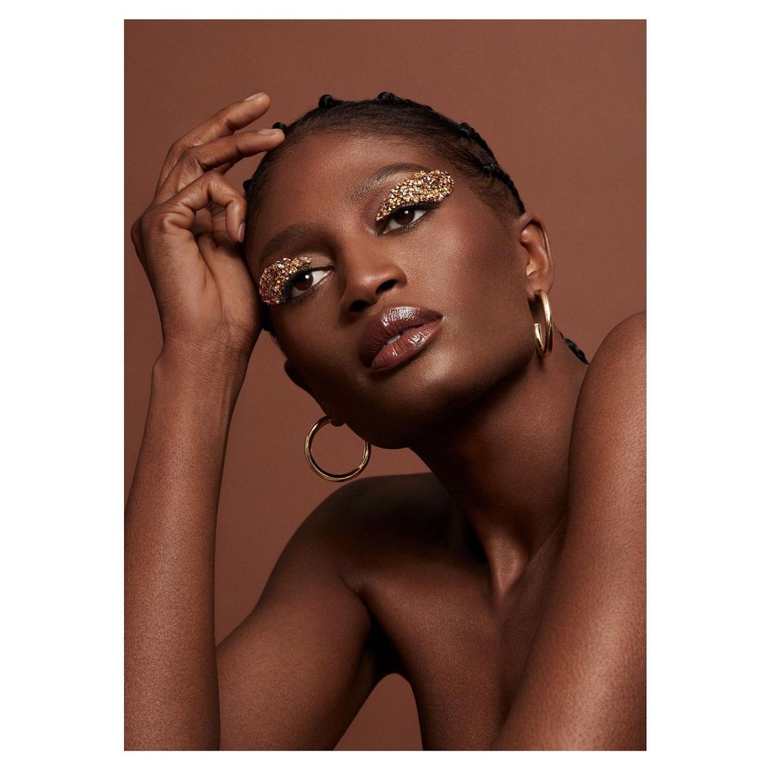 Beauty shot of an African American female model with glittery eyeshadow, gold hoop earrings, glossy lips, and braided hair against brown backdrop.