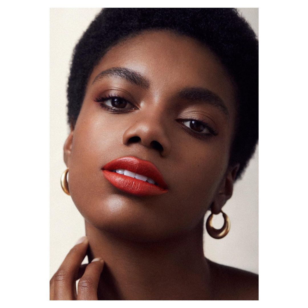 Close-up beauty portrait of a Black woman with short dark hair wearing bold red lipstick and gold hoop earrings.