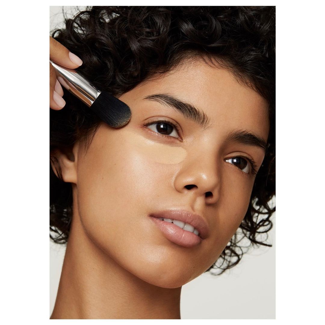 Close-up of a young woman applying makeup with a brush, showcasing a beauty routine in a bright, soft light setting.