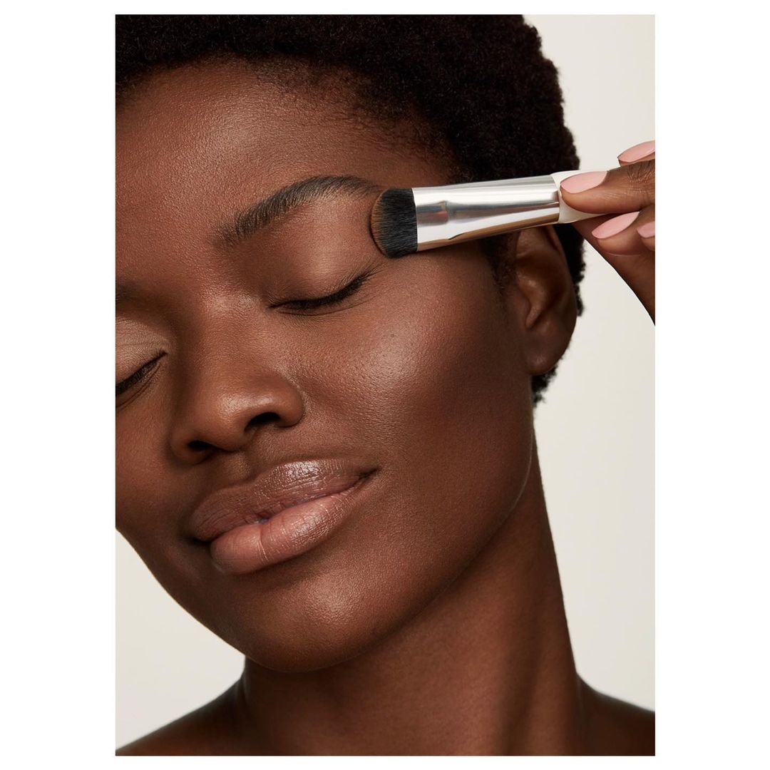 Close-up of a woman with dark skin and short hair applying makeup to her eye with a brush against a light-beige background.