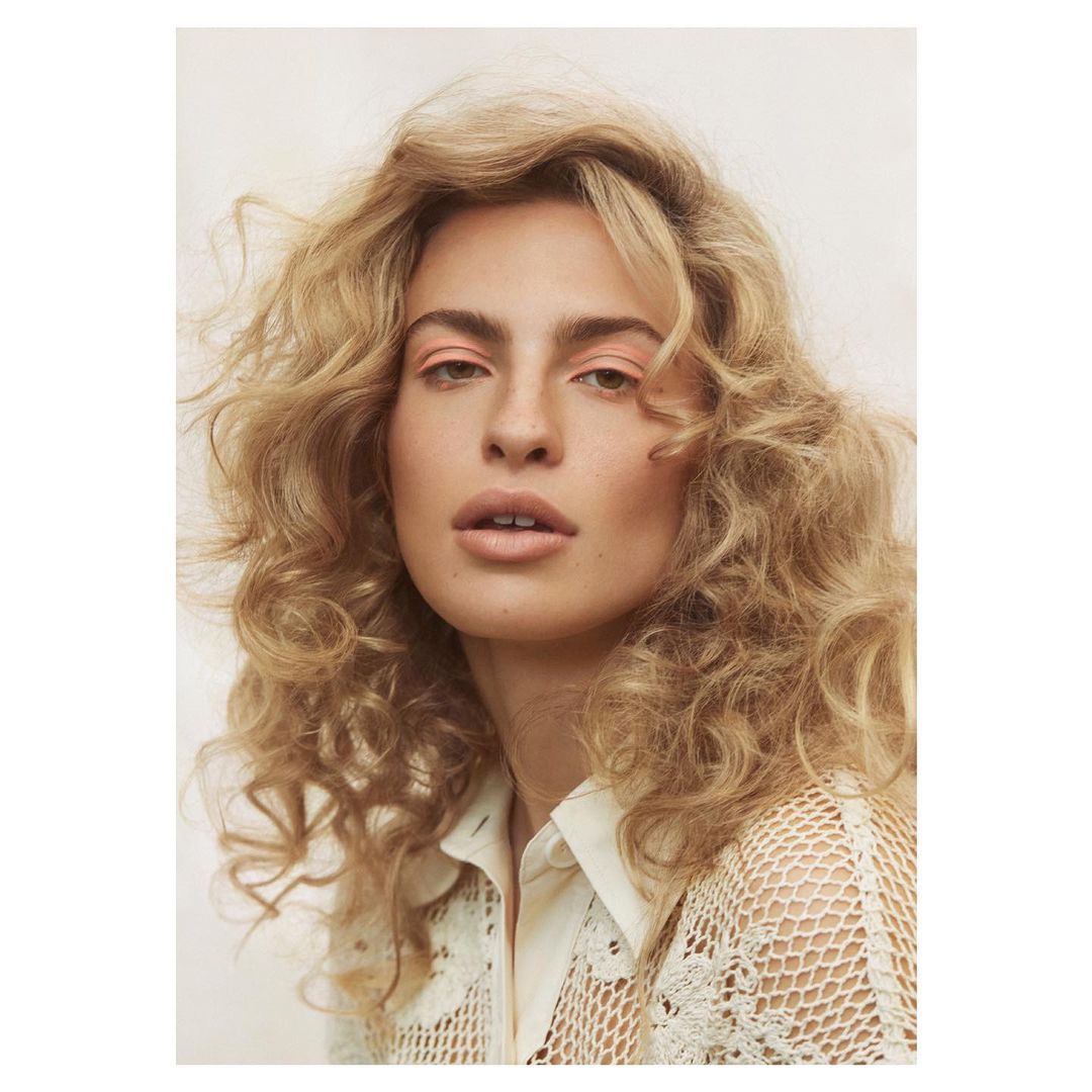 Woman with blonde curly hair poses in a studio with a cream-colored crochet shirt. Light and airy fashion and beauty photography.