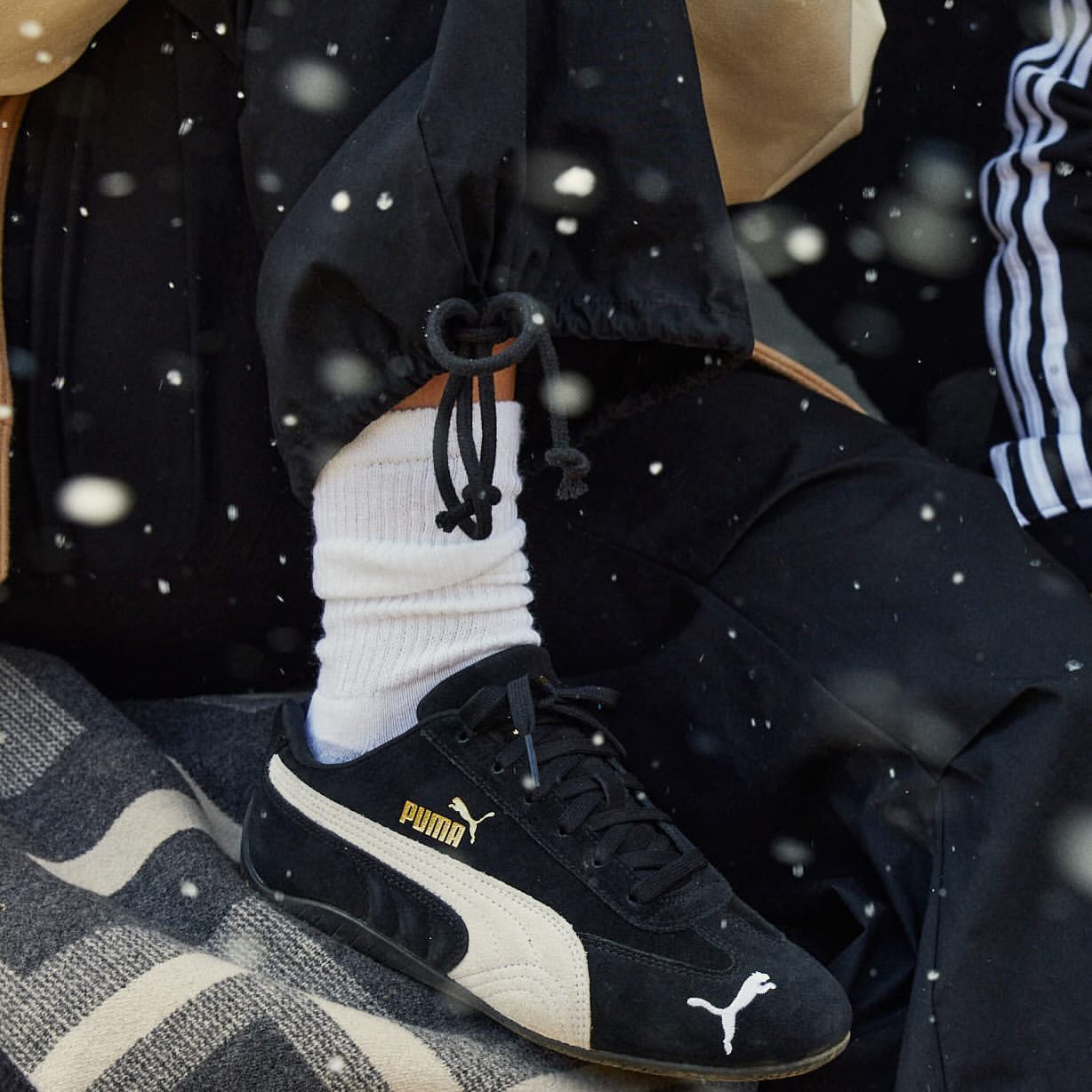 Close-up of a person wearing black Puma sneakers with white socks and black trousers, shot by Nick Eucker.