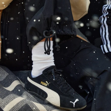 Close-up of a person wearing black Puma sneakers with white socks and black trousers, shot by Nick Eucker.