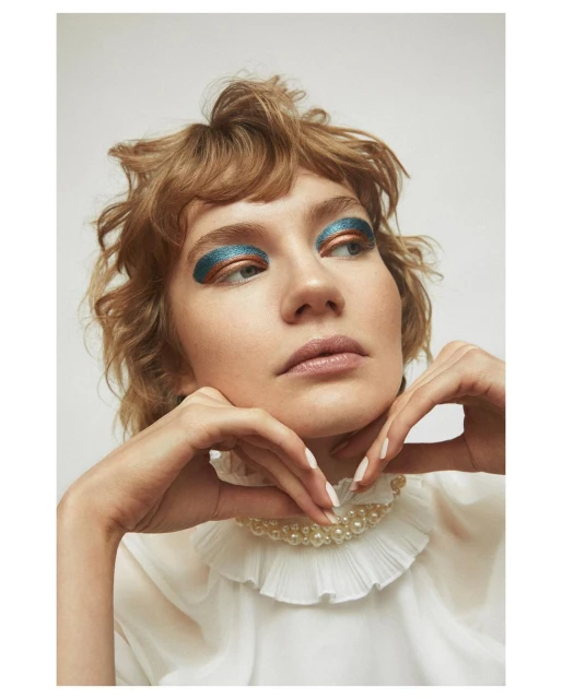Close-up studio portrait of a blonde woman with creative teal and copper eyeshadow, wearing a white ruffled top and pearl necklace.