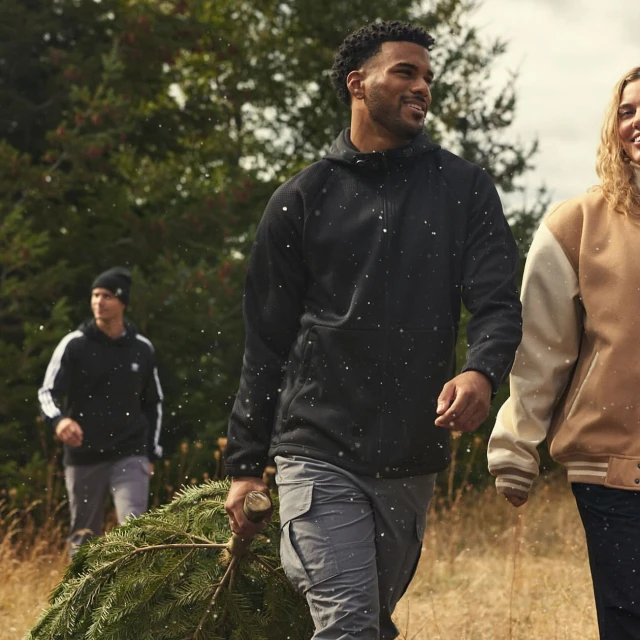 Three people walking with a Christmas tree through a grassy field with trees in the background, wearing casual winter clothing.