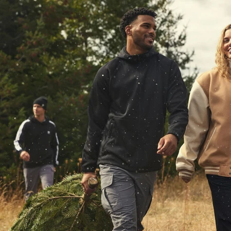 Three people walking with a Christmas tree through a grassy field with trees in the background, wearing casual winter clothing.