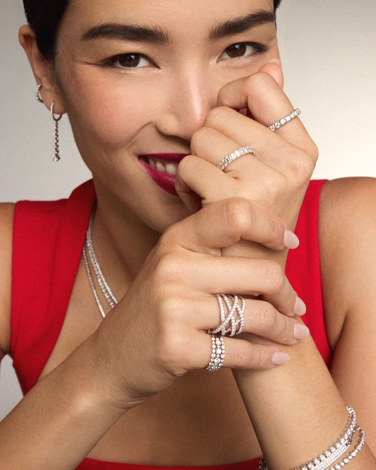 Close-up of a smiling woman with dark hair and red lipstick, wearing a red top and several diamond rings, earrings, and necklaces.