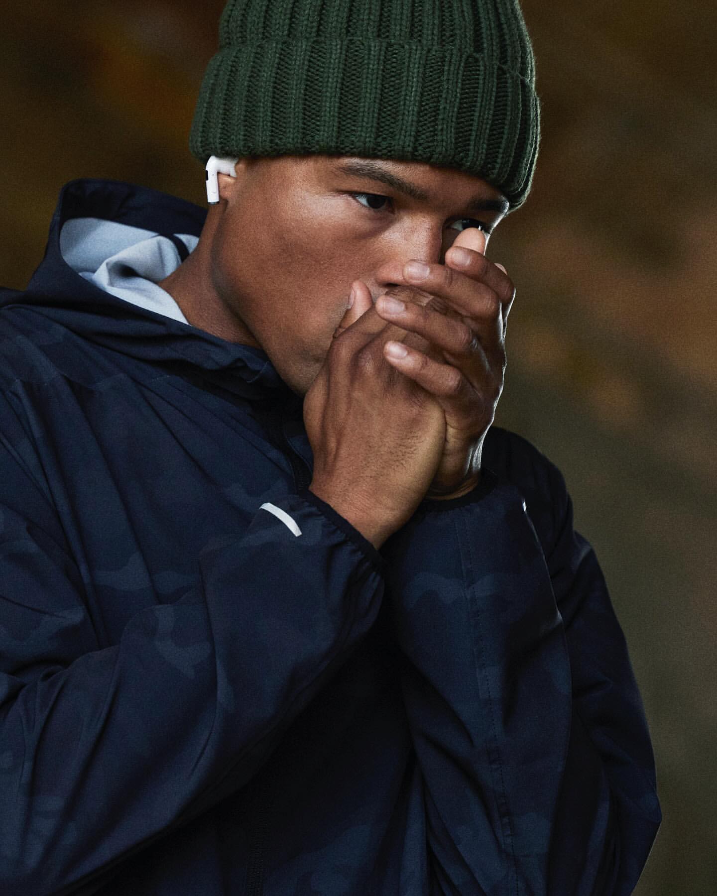 Close-up of a man wearing a beanie and hooded jacket, warming his hands by blowing on them, set against a neutral background.