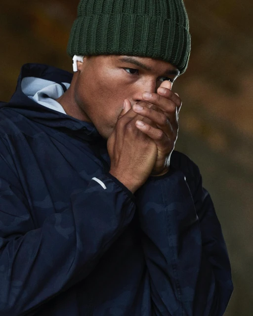 Close-up of a man wearing a beanie and hooded jacket, warming his hands by blowing on them, set against a neutral background.